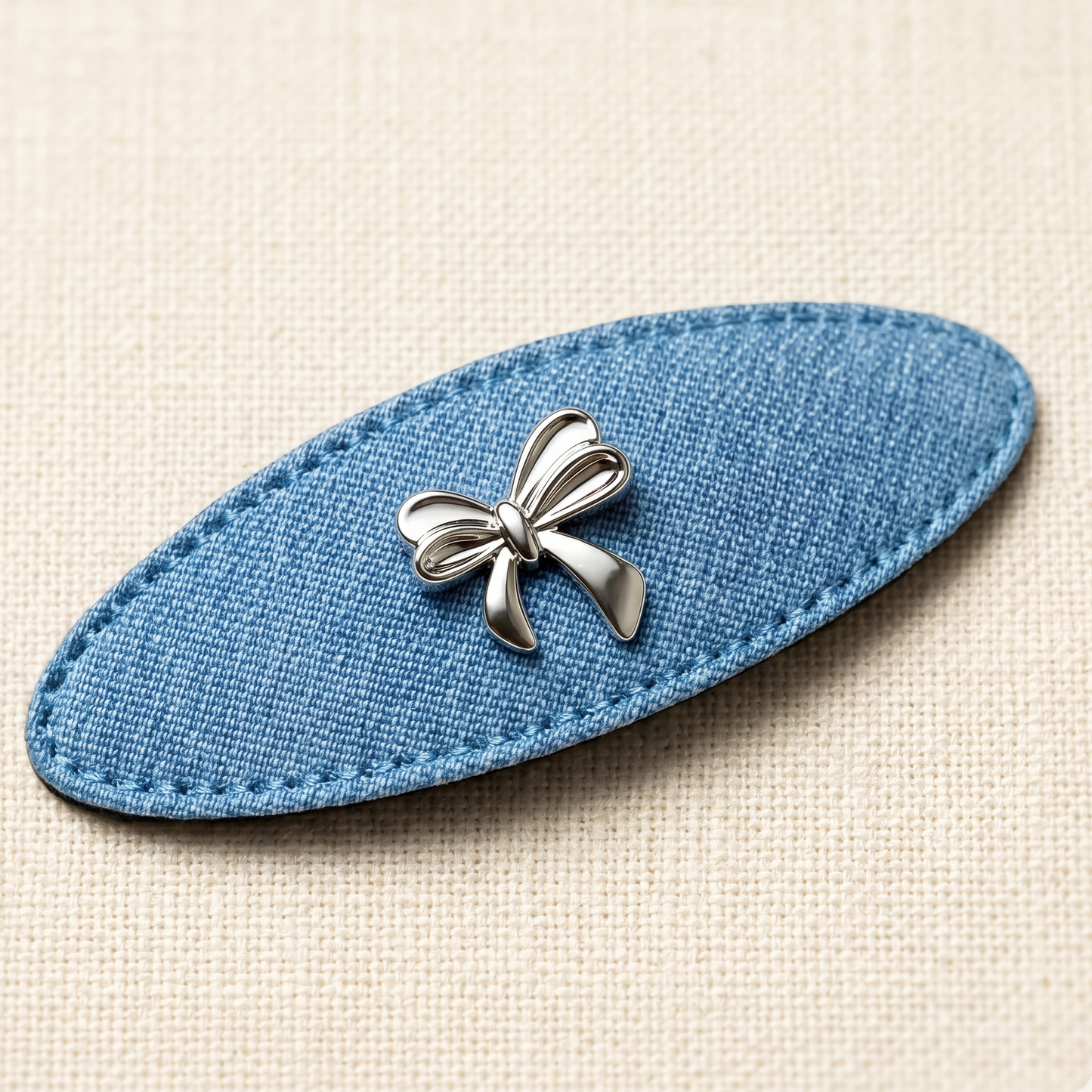 Blue oval fabric hair clip with a silver bow design on a beige background